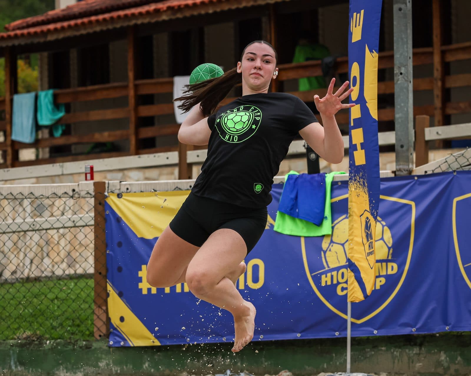 Atleta saltando para arremessar na areia, bola verde na mao e mata ao fundo