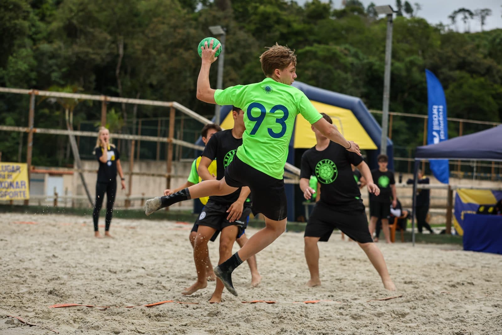 Disputa de bola no jogo de areia, atletas de colete verde e preto no salto